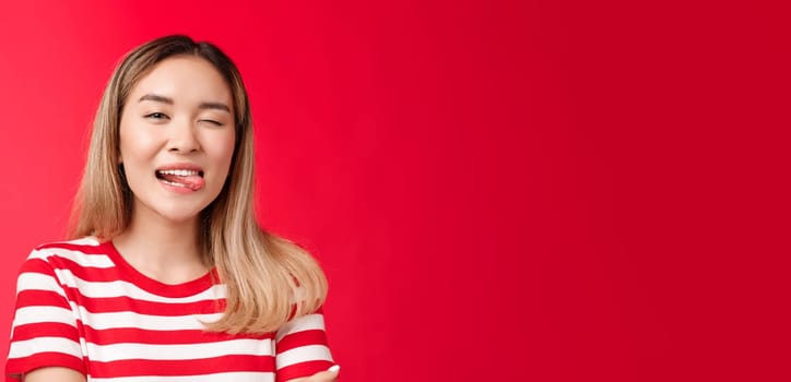 Close-up sassy carefree outgoing urban asian female student enjoy summer vacation wear striped t-shirt, show tongue playfully winking camera, stand red background positive friendly mood.
