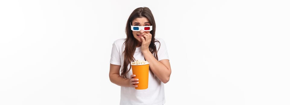 Entertainment, fun and holidays concept. Portrait of scared young cute girl in 3d glasses, eating popcorn, stooping and biting fingers as being scared of horror movie watching in cinema.