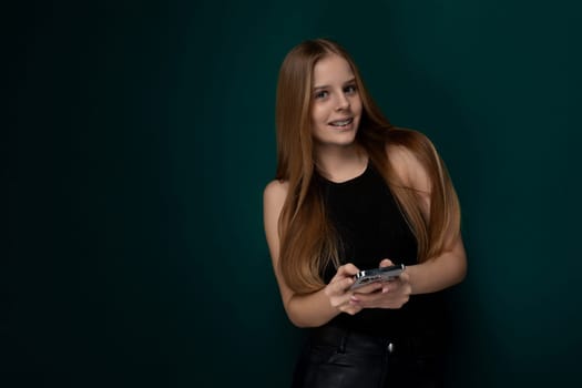 A woman with vibrant long red hair is holding a modern cell phone in her hand. She appears focused on the screen, possibly texting or scrolling through social media. The background is nondescript, allowing the viewer to focus on the woman and her phone.