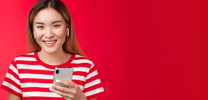 Girl staying tuned watching favorite vloggers wear wireless earphones, smiling joyfully camera toothy white grin, hold smartphone, pick song, browsing music app platform, red background.