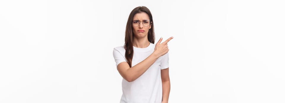 Waist-up portrait of moody, disappointed and complaining cute brunette woman in glasses, frowning and grimacing, sulking defensive, pointing finger upper left corner at something offensive.