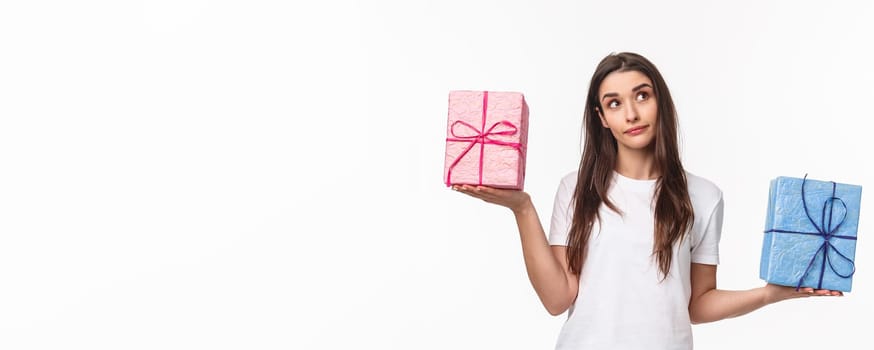 Celebration, holidays and presents concept. Portrait of thoughtful young girl taking decision, look up wondered, weighing gift boxes in hands spread sideways, what is heavier, white background.