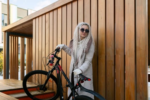 A woman stands next to a bike parked by a weathered wooden wall. She is dressed casually and appears to be taking a break from cycling.