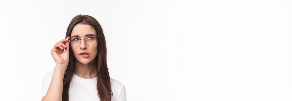 Close-up portrait of smart and thoughtful beautiful young girl in glasses, thinking, squinting suspicious and biting lip look up while pondering something, have interesting idea, white background.