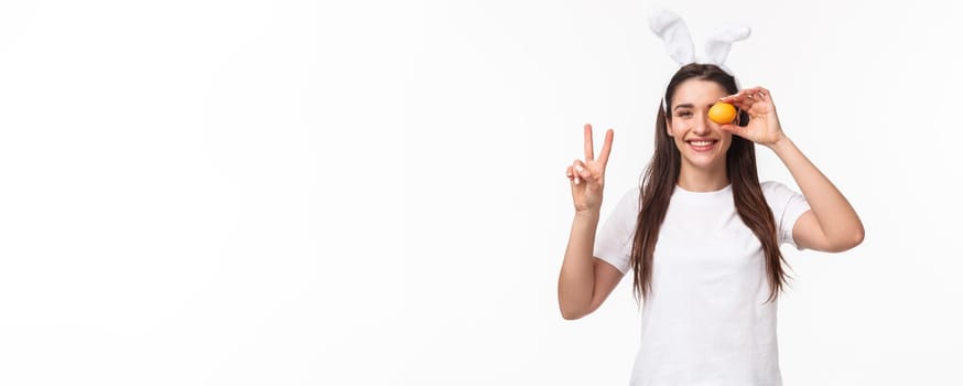 Portrait of peaceful and cheerful young cute female in white t-shirt, showint peace sign and hold colored egg over eye, smiling happy, enjoying traditional holiday celebration, Easter day.