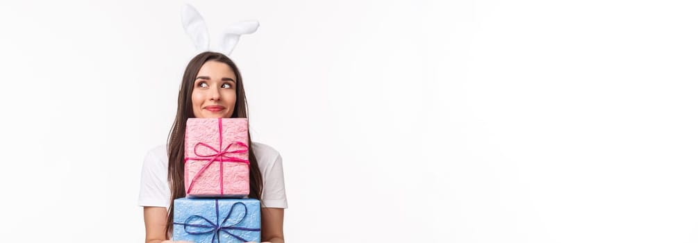 Celebration, holidays and presents concept. Portrait of silly and cute, lovely young girl in rabbit ears, holding presents, daydreaming, smiling and looking away, giving out gift, white background.