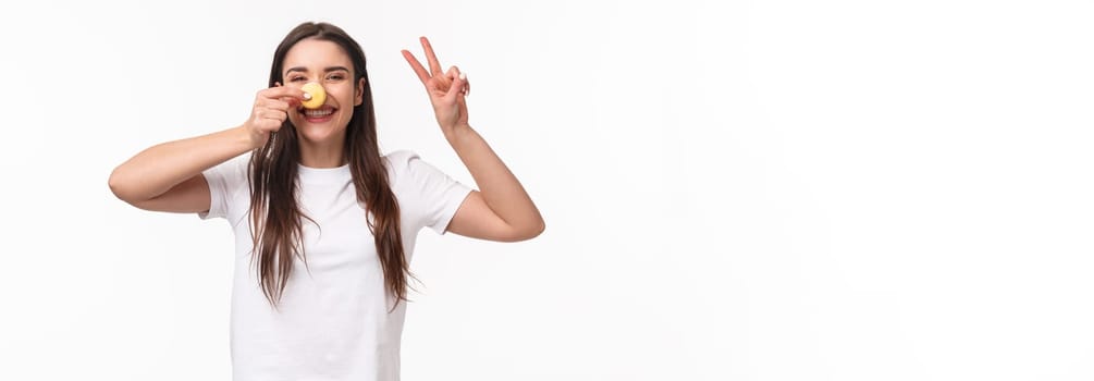 Portrait of positive, cheerful young girl eating sweets, holding macaron over nose, make peace sign, look kawaii and silly, laughing joyful, visit favorite dessert cafe, white background.