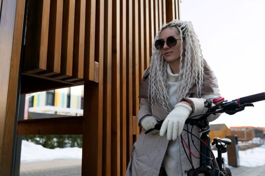 A woman is standing next to a black bike in front of a modern building. She is wearing casual clothing and seems to be taking a break from riding. The architecture of the building is contemporary with large windows and a sleek design.