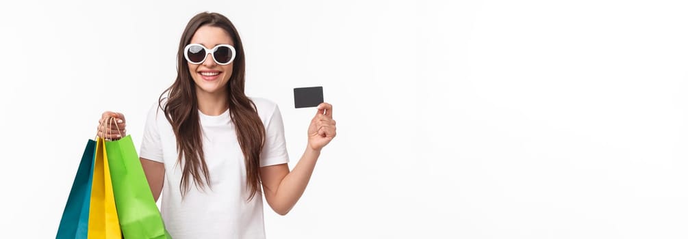 Shopping, leisure and lifestyle concept. Portrait of carefree, relaxed young woman spending her money on purchasing new clothes, products, holding shop bags and showing credit card, smiling pleased.