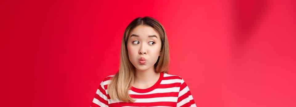 Close-up silly tender young modern asian girl blond haircut hold breath, folding lips and pouting look away as if hiding secretn, stand red background innocent clueless seal lips with promise.