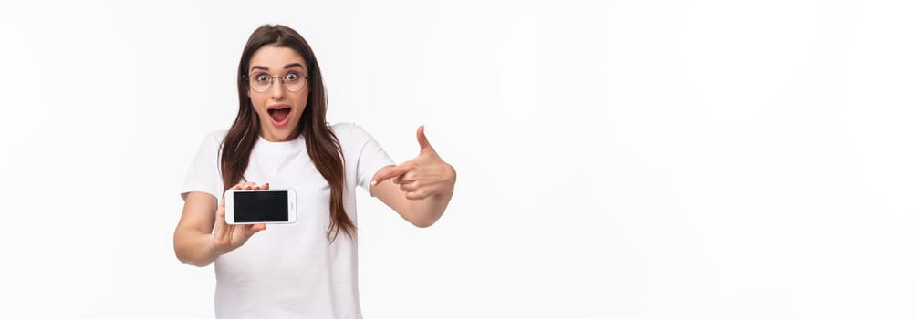 Communication, technology and lifestyle concept. Portrait of surprised and excited young woman in glasses hurry up with announcement of awesome new application, pointing at mobile phone.