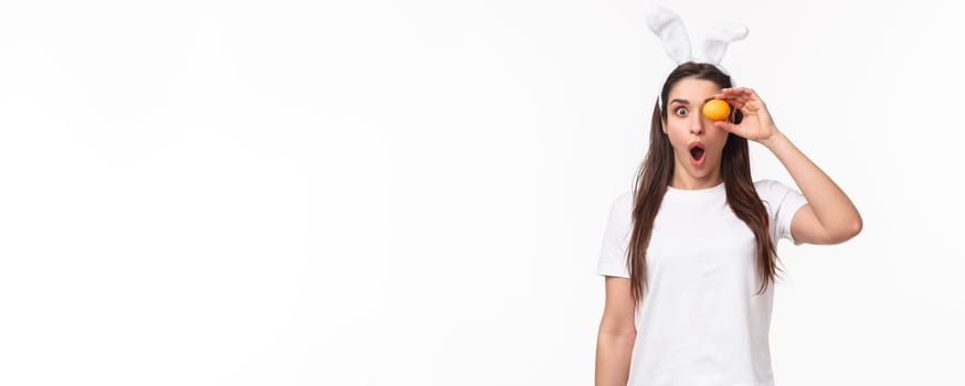 Waist-up portrait of amazed and surprised young woman seeing something awesome, hold colored egg over eye, staring shocked camera, gasping impressed, wear rabbit ears, celebrate Easter day.