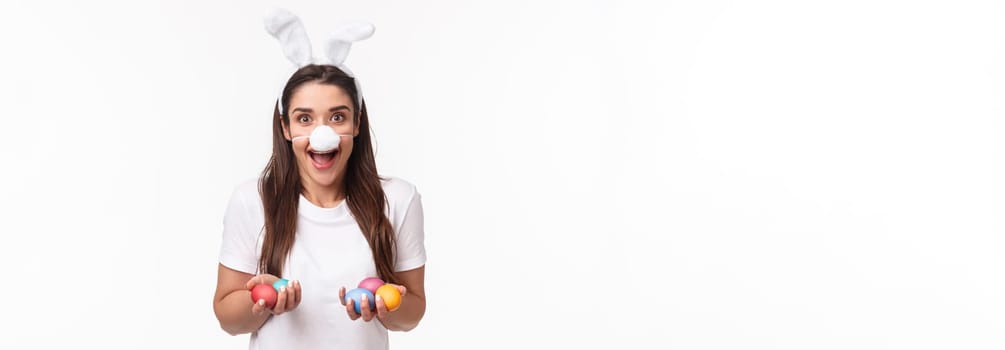 Portrait of excited, happy and carefree young woman spending holiday with family, wearing funny rabbit ears and nose, holding colored eggs, painting it with kids on Easter day, white background.