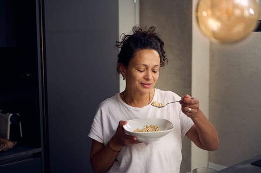 Young woman in white t-shirt, enjoying her wholesome breakfast, eating a bowl of muesli, standing at kitchen counter at home. People. Food consumerism. Diet and slimming concept. Healthy lifestyle.