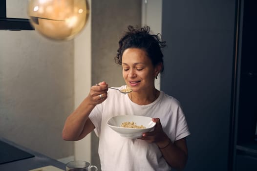 Latin American young woman 40s, smiling, eating healthy muesli for breakfast at home kitchen, dressed in white pajamas