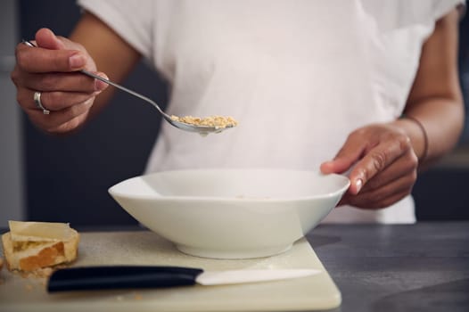 Details on the hand of woman in white t-shirt, holding a spoon with wholegrain muesli granola above a white bowl, enjoying a healthy breakfast in the morning. Healthy eating, slimming and diet concept