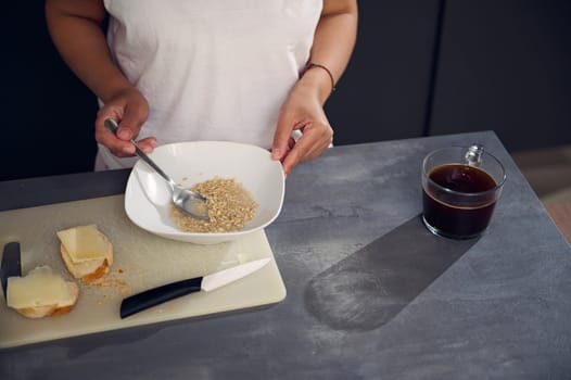 Top view woman in white pajamas, holding a spoon with wholegrain muesli granola above a bowl, enjoying a healthy breakfast in the morning, standing at kitchen table with a cup of aroma coffee