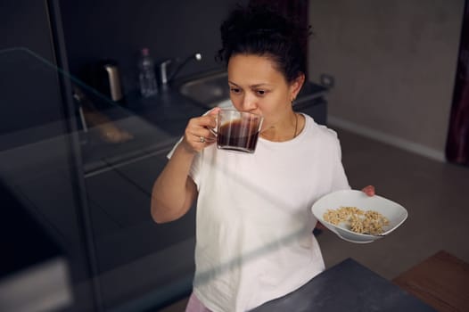 Beautiful young woman having a breakfast before work. Multi ethnic lady in pajamas eating oats with vegetarian milk and drinking freshly brewed espresso coffee. Start your day with healthy breakfast