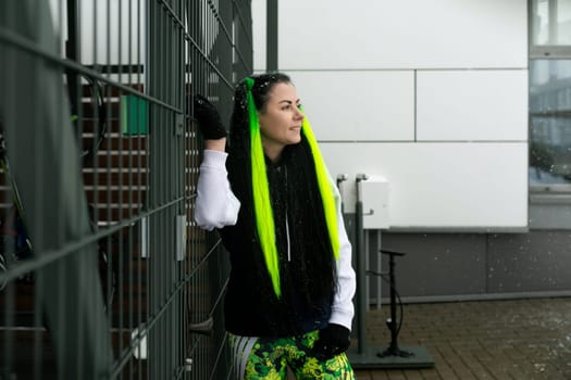 A woman with striking bright green hair stands confidently next to a weathered wooden fence, the sun casting shadows across the scene.