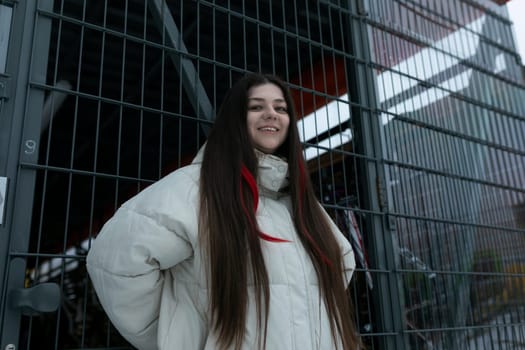 A woman with long hair is standing in front of a wooden fence. She looks at the camera confidently with an unassuming expression. The scene captures her in a casual moment, surrounded by the rustic charm of the fence.