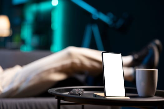 Focus on mockup device with man sleeping on couch in blurry background in neon lit apartment. Close up of isolated screen smartphone on coffee table in front of person snoozing