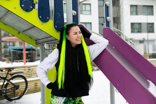 A woman with long hair is standing next to a vibrant purple and green bench in an urban setting. She appears relaxed and is looking off into the distance.