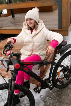 A woman is seen sitting on a bench next to a bicycle. She appears relaxed and is looking around. The bike is parked beside her, leaning against the bench.