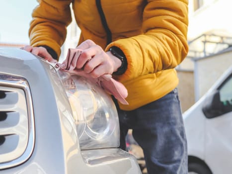 A young unrecognizable Caucasian brunette in a yellow jacket, bending over, polishes the headlight of a gray car with a pink rag on a city street in front of the house, close-up view from below. The concept of home car wash, headlight polishing.