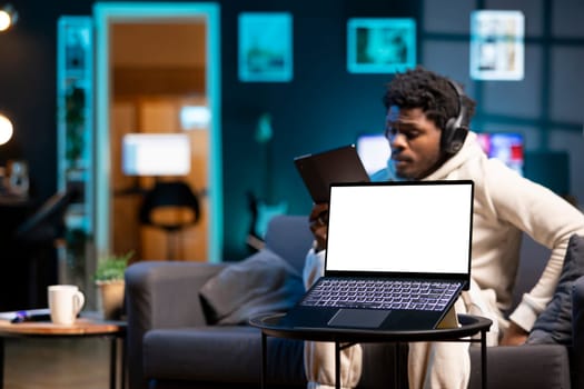 African american man taking break from work to listen to music and read from tablet, focus on mockup laptop. Teleworker wearing headphones, enjoying leisure time next to isolated screen notebook