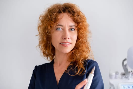 Closeup portrait of female cosmetologist at medical office. Redhead woman dermatologist wearing scrubs holding tool at hospital. Skincare practitioner in medical uniform in aesthetic clinic office.