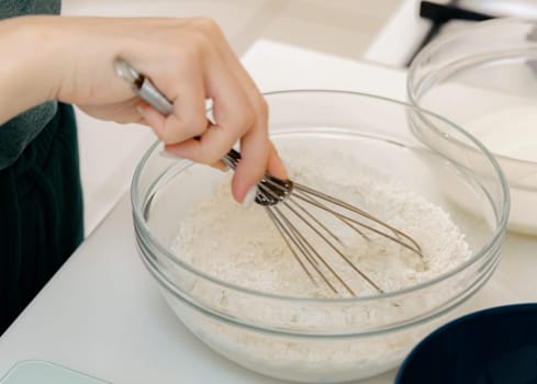 The hand of one young Caucasian unrecognizable girl mixes the dough manually with a whisk in a glass bowl, close-up on the side. Step-by-step instructions for baking synabons. Step 2.