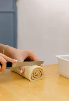 One unrecognizable young Caucasian girl cuts dough with brown sugar with cinnamon into pieces using a knife on a wooden table, standing in the kitchen, close-up side view. Step-by-step instructions for baking synabons. Step 9.