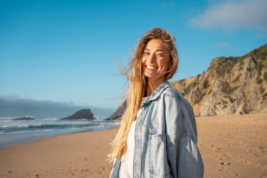 Portrait of smiling young woman with long blond hair on sandy beach. Happy female in denim shirt with sea and mountain in background. Girl enjoying vacation against blue sky.