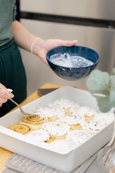 One unrecognizable young Caucasian girl spreads white cream on round hot buns with brown sugar and cinnamon in a white baking dish on a wooden table, standing in the kitchen, close-up top view. Step-by-step instructions for baking synabons. Step 13