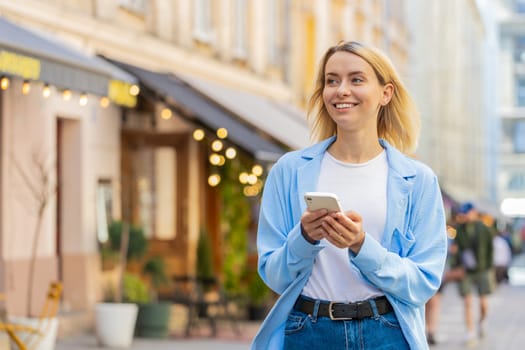 Caucasian young woman using smartphone typing texting social media messages e-mail looking for a way on map in mobile navigator app outdoors. Smiling traveler walking on city street. Town lifestyles.