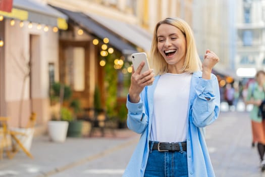 Caucasian young woman using mobile smartphone celebrating win good message news, lottery jackpot victory, giveaway online outdoors. Happy adult lady tourist standing in city street. Town lifestyles