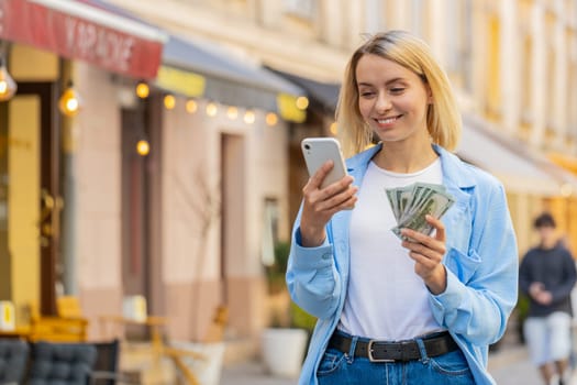 Happy rich young blonde woman counting money dollar cash, use smartphone calculator app, plans to order gifts and food delivery online, booking hotel room outdoors. Lady standing on urban city street.