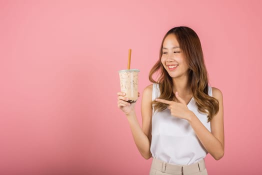 Pearl milk tea beverage concept. Asian beautiful young woman holding drinking brown sugar flavored tapioca pearl bubble milk tea and point finger, smile portrait female, isolated on pink background