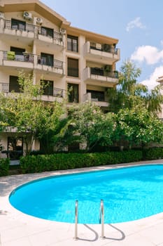 Oval swimming pool among flowering bushes and green trees in the courtyard of a multi-storey hotel. High quality photo