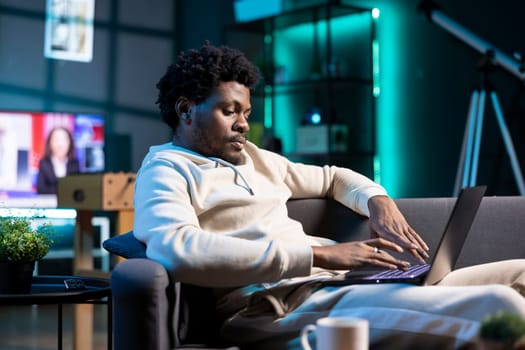 Relaxed remote worker checking his emails on laptop while laying on couch. BIPOC freelancer working from home in bright stylish cozy apartment personal office, typing on notebook