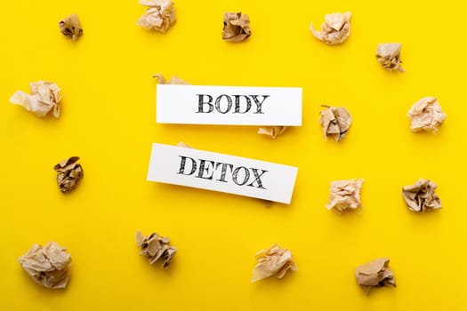 Two pieces of paper with the words body detox written on them. The paper is placed on top of a yellow background, which is covered in crumpled paper. Scene is somewhat chaotic
