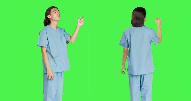 Asian medical assistant sees hologram in studio with greenscreen layout, working with holographic image and artificial intelligence. Male nurse in uniform with healthcare expertise.