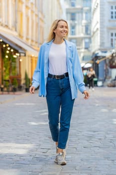 Portrait of young blonde tourist woman walking on urban city street outdoors. Smiling happy lady girl traveler having positive good mood enjoying in summer daytime. Town lifestyles, vacation holidays