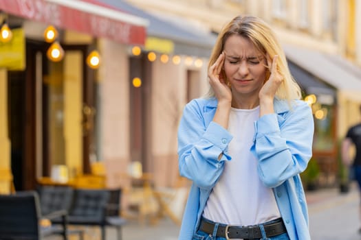 Displeased exhausted Caucasian woman rubbing temples to cure headache problem suffering from tension migraine stress. Young lady tourist grimacing pain high blood pressure stands on city urban street