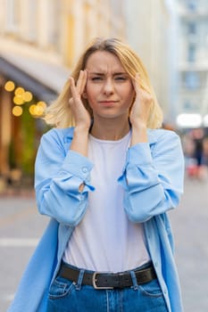 Displeased exhausted Caucasian woman rubbing temples to cure headache problem suffering from tension migraine stress. Young lady tourist grimacing pain high blood pressure stands on city urban street