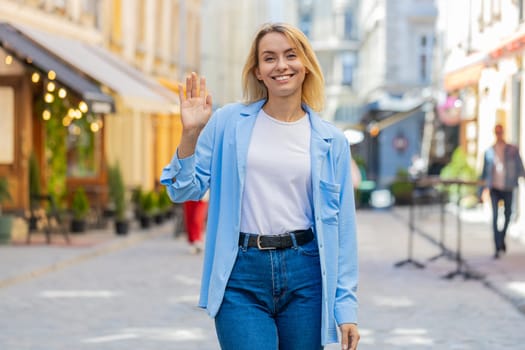 Cheerful young woman tourist smiling friendly at camera, waving hands gesturing invitation hello, hi, greeting, goodbye, welcoming with hospitable expression outdoors. Lady on urban city town street