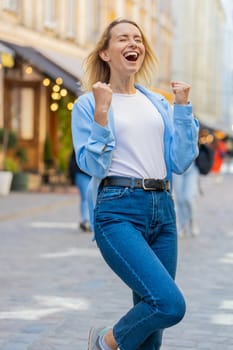 Excited happy woman traveler shouting, celebrating success, winning, goal achievement, good news, lottery jackpot luck, victory outdoors. Young lady walking on urban city sunshine street. Lifestyles