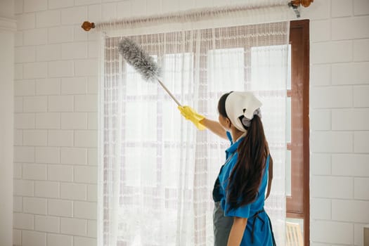 Asian woman smiles dusting window blinds with a duster. Her routine cleaning ensures a modern clean home. Portrait of an adult occupied with household hygiene and purity. whisk