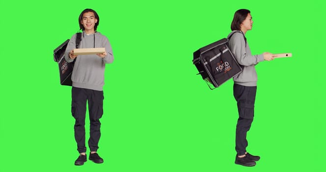 Asian man delivering food for clients using thermal bag over full body greenscreen, carrying meal orders. Deliveryman employee posing on camera, providing takeaway aliments.