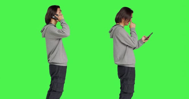 Young man answering call and texting in studio with greenscreen backdrop, talking remotely on smartphone and messaging. Asian male model using mobile phone for different actions.